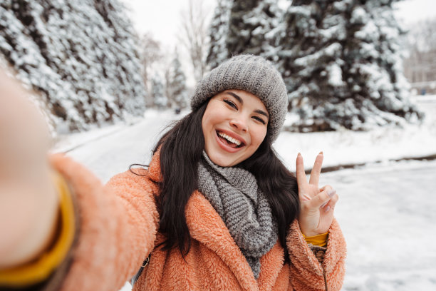 在冬天寒冷的日子里，戴着灰色帽子和粉色外套的漂亮快乐的女人在自拍和玩猿。女孩在冬天自拍图片下载