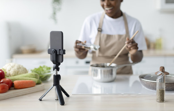 满意的千禧黑人女士在围裙做饭在家里通过电话在厨房内部拍摄视频课程，剪裁图片下载