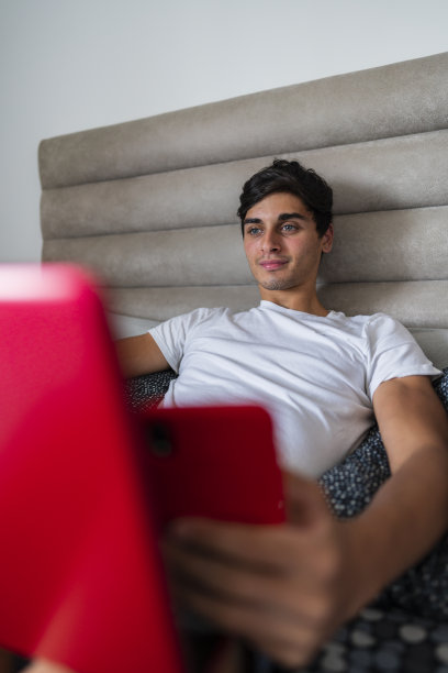 一个年轻的拉丁美洲人坐在家里房间的床上，手里拿着手机查看他的银行信息，用笔记本电脑确认这些信息图片下载