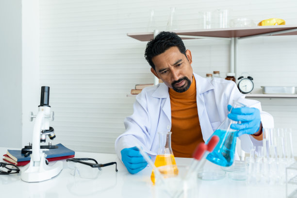 男老师，穿着白色实验服，戴着橡胶手套，架子和桌子上有许多实验工具，抓起桌子上的蓝色试剂瓶仔细检查。在开始化学课之前图片下载