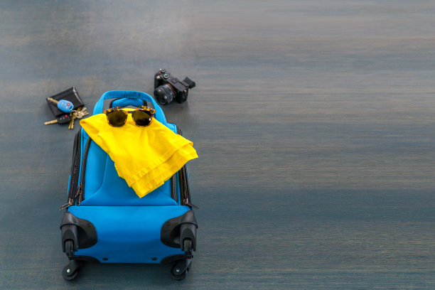 蓝色旅行袋，里面有黄色衬衫，太阳镜，钱包，钥匙和相机，放在灰色的水泥地上。准备去度假。图片下载