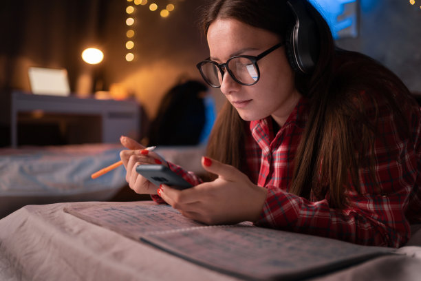 女孩戴着耳机，躺在床上用手机学习。可爱的女学生使用智能手机技术，在宿舍的卧室里听课程，看视频课程，用手机在线学习图片下载