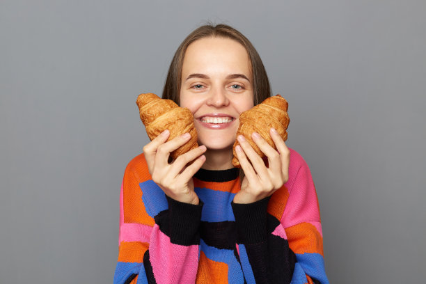 室内拍摄的快乐的年轻成年白人女子穿着毛衣摆出孤立的灰色背景，对着镜头微笑，展示美味的羊角面包。图片下载
