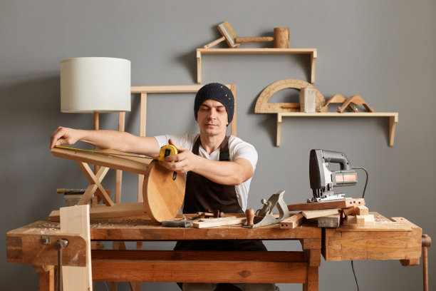 严肃英俊的白人细木工肖像，戴着围裙和帽子坐在木工车间的桌子旁，测量他的车间制作的椅子腿的长度。图片下载