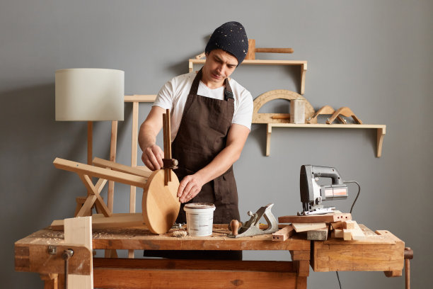 穿着棕色围裙和黑色帽子的木匠，在车库或工作室用特制的仪器手工测量木材的厚度，在木工车间制作椅子的工匠。图片下载