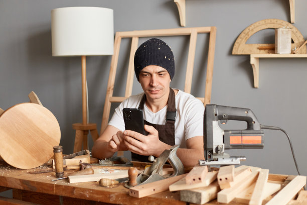 室内镜头，一名工匠坐在他的车间工作区域的桌子旁，用手机发信息或阅读新闻，搜索有关木工的信息。图片下载