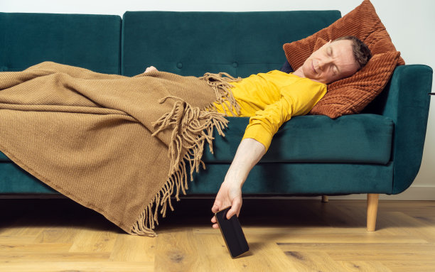 疲惫的男人睡在沙发上，手拿着智能手机。过度工作的人躺在枕头上，盖着毯子。图片下载