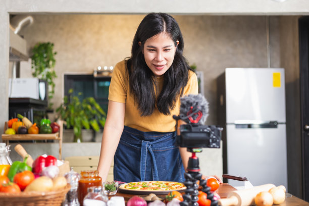 年轻迷人的亚洲女子在网上直播中交流如何在室内厨房烹饪自制披萨。内容创作者使用相机记录食品审查。图片下载