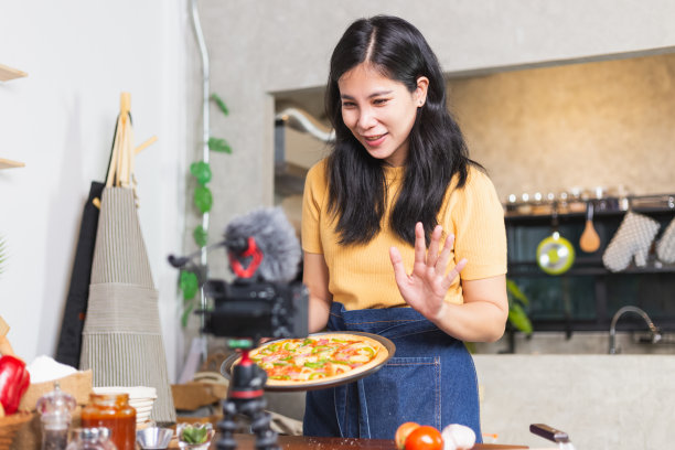 年轻迷人的亚洲女子在网上直播中交流如何在室内厨房烹饪自制披萨。内容创作者使用相机记录食品审查。图片下载