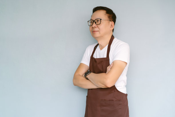 侧视图中年亚洲商人穿着围裙折叠手臂，看着远处思考未来的职业机会，微笑着站在绿色粉彩墙与复制空间。图片下载