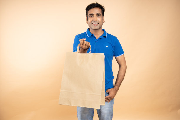 快乐的年轻印度男子穿着休闲t恤拿着棕色纸购物袋站在米色工作室背景孤立。电子商务销售和优惠。模型图片下载