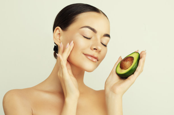 美丽女人的肖像，她手里拿着半个牛油果。孤立的浅米色工作室背景。图片下载