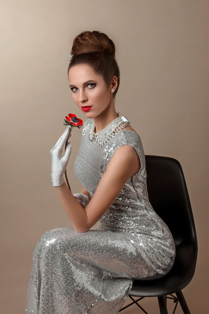 一位年轻漂亮的黑发女子，穿着闪亮的白色连衣裙，坐在一把黑色的椅子上。一位时髦的模特手里拿着一朵红花。图片下载