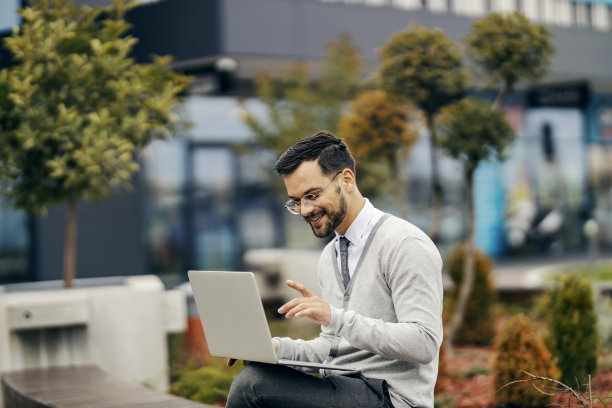 一位穿着时髦休闲装的年轻快乐的商人坐在公园外面，一边对着笔记本电脑微笑一边打字。图片下载