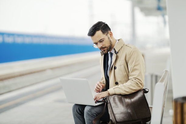 一个忙碌的年轻绅士坐在火车站的长凳上，一边等火车一边用笔记本电脑打字。图片下载