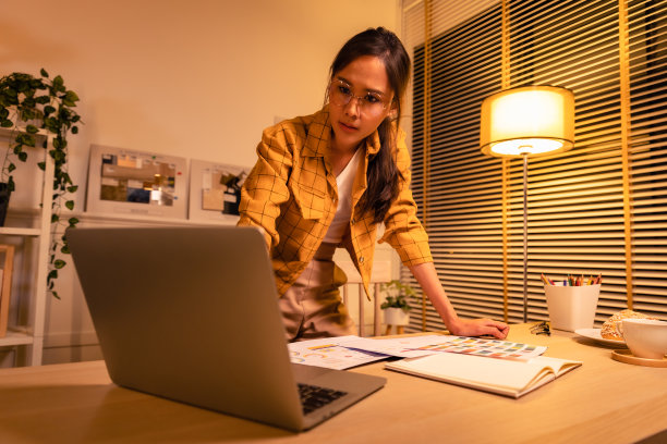 年轻漂亮的女人在黄色衬衫工作到深夜从家里在客厅加班。女商人在工作场所低头看电脑。图片下载