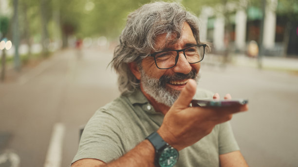 阴沉沉的，满脸笑容的中年男子，头发和胡子都花白了，坐在长凳上用手机。戴眼镜的成熟绅士在免提上录制语音识别信息图片下载