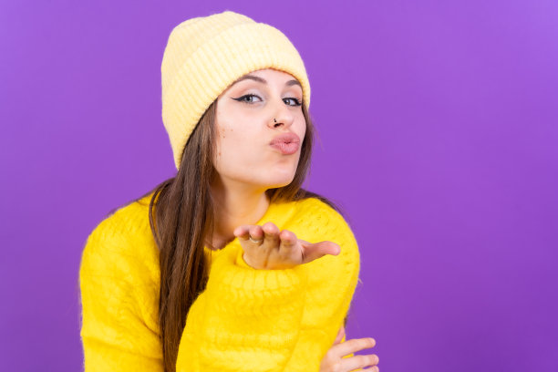 照片肖像的年轻女子吹一个吻孤立在紫色背景，黄色羊毛衫图片下载