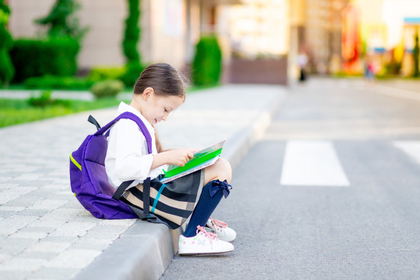 一个一年级的女孩背着书包，手里拿着一本书在学校读书或做作业，这个概念就是回到学校图片下载