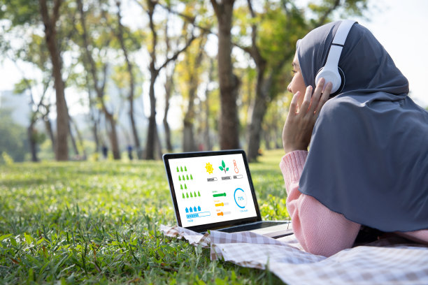 一名穆斯林妇女在她屏幕上的信息图表上学习了生长植物后，用笔记本电脑在公园的草地上听音乐。自由职业者，女性工作者，环境和地球概念的教育家图片下载