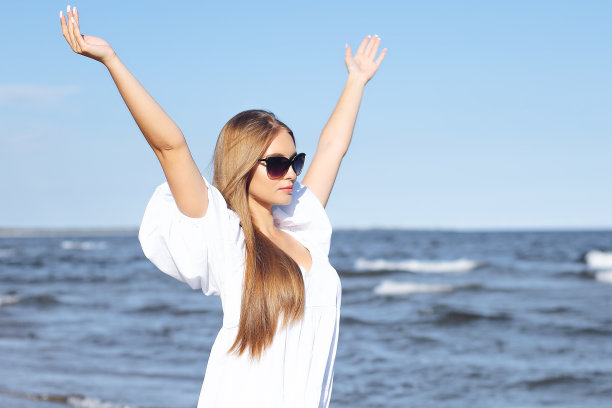 快乐的金发女人在海滩上，穿着白色连衣裙，戴着太阳镜，举手图片下载