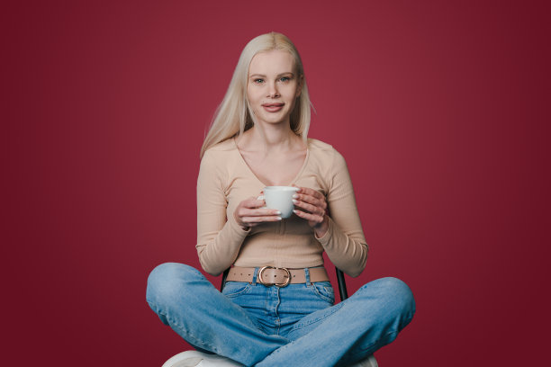 美丽积极的女人坐在椅子上拿着一杯咖啡，微笑着在深红色的背景上孤立的相机。迷人的漂亮女孩。人们的生活方式。图片下载