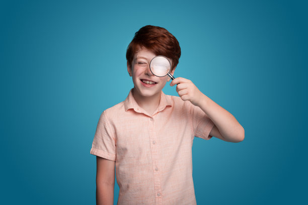 穿着休闲装的男生站在蓝色背景下，通过放大镜看着相机。教育的概念。搜索概念。孤立的肖像。学校教育。图片下载