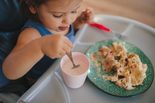 有趣的小女婴从绿色盘子里拿着勺子在她的杯牛奶的肖像。儿童发展技能概念。图片下载