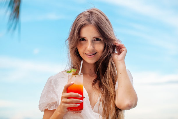 迷人的年轻女子在白色连衣裙品味鸡尾酒在海滩上图片下载