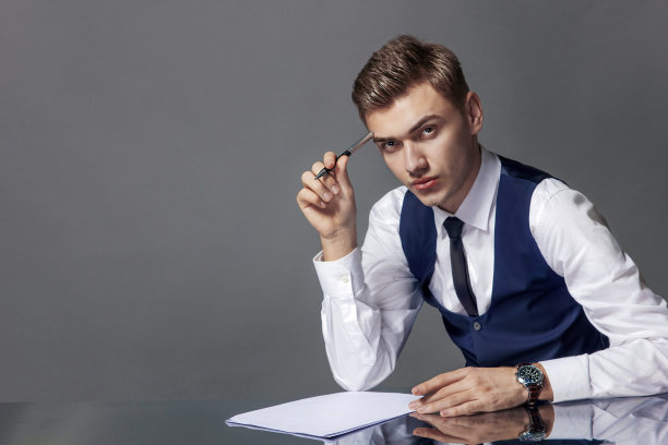 一个西装革履的男人坐在办公桌前，手里拿着一支笔，放在灰色背景上的一张白纸上。图片下载