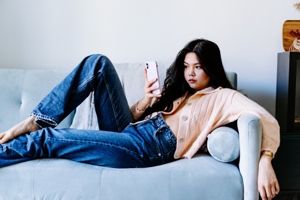 年轻的亚洲女性戴着耳机听音乐，在沙发上跳着充满活力和节奏的舞蹈。迷人的亚洲女孩看着智能手机坐在客厅里度过闲暇时间在家里图片下载