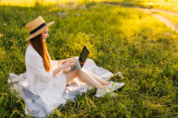 愉快的红头发妇女在草帽使用笔记本电脑坐在绿色的草地在夏季公园。漂亮的女士购物或在社交网络上在线聊天。图片下载