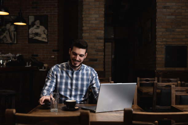 男性自由职业者坐在餐厅的桌子旁，喝着咖啡，用笔记本电脑工作。外籍男子在自助餐厅在互联网上做研究和全球业务图片下载