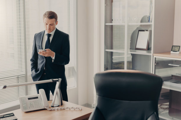 帅气的年轻男经理，穿着公司服装，用智能手机浏览网页和在线聊天，目光集中，在现代办公室内部摆姿势，阅读新闻和浏览信息图片下载