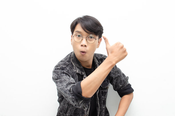 在白色的背景下，一位穿着牛仔裤夹克的帅气迷人的亚洲男子正开心地笑着，竖起大拇指表示赞赏。男人通过竖起大拇指来表示赞同和赞美。图片下载