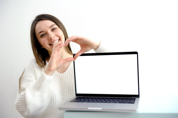 白色背景的女孩指着笔记本电脑屏幕，白衣女孩在笔记本电脑旁边用手画了一颗心，微笑着，笔记本电脑上有空白的地方可以放置任何内容的广告文字图片下载