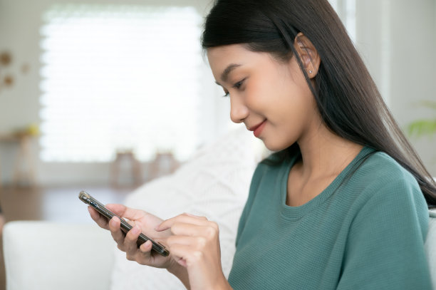 快乐的年轻亚洲女子在家里放松，她坐在沙发上，用手机图片下载