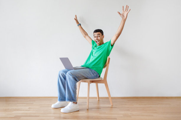 快乐的阿拉伯男子坐在椅子上，用笔记本电脑高兴地举起双手，在网上收到好消息，有复制空间图片下载