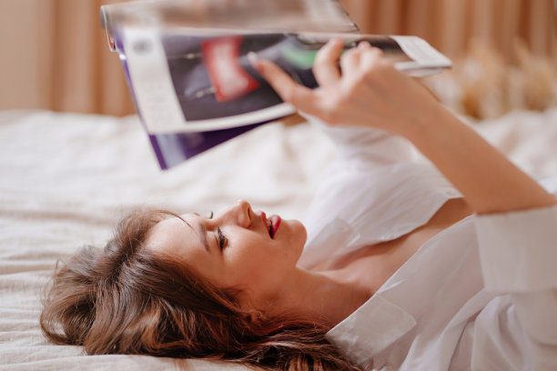 一个迷人而快乐的女人躺在床上看杂志。图片下载