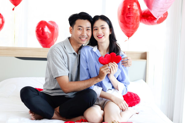 幸福的亚洲情侣抱着红心，坐在卧室的白色床上互相拥抱，浪漫的恋人在情人节庆祝难忘的纪念日，爱的庆祝概念。图片下载