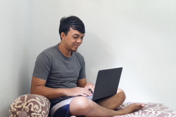 肖像亚洲眼镜男子正微笑着看着笔记本电脑在卧室的工作与自然光线的概念图片下载