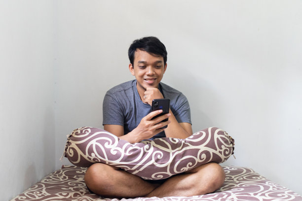 一名亚洲男子在一间有自然光的卧室里微笑着看智能手机图片下载