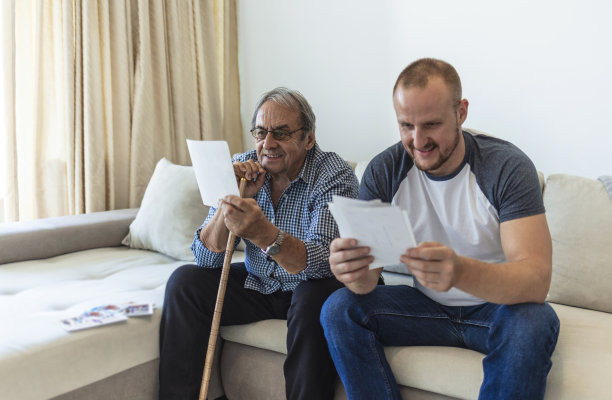 一个成年的时髦儿子和年长的父亲坐在家里的楼梯上，看着照片。年长的父亲和成年的儿子在家里聊天和看照片。图片下载
