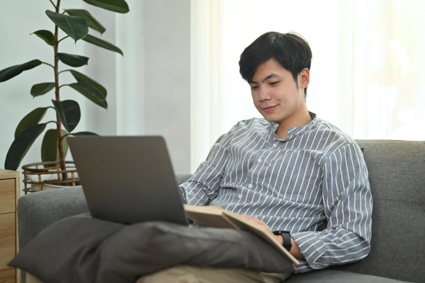 愉快的年轻亚洲男子上网，在笔记本电脑上检查电子邮件，而坐在舒适的客厅。图片下载