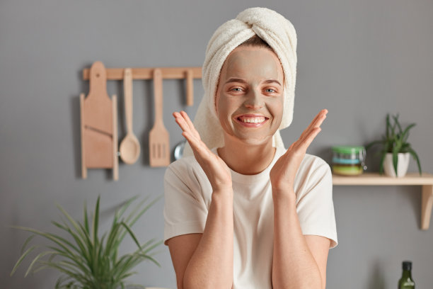 有吸引力的迷人的白人妇女与粘土面膜的脸皮肤和被毛巾包裹在厨房里，享受早晨的皮肤护理程序，保持手掌靠近脸。图片下载