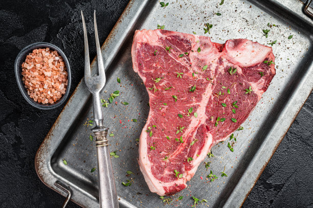 t骨牛排，生的大理石纹牛肉，放在厨房的钢托盘里，配上香草。黑色背景。俯视图图片下载