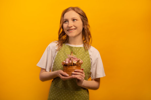 快乐的微笑梦幻的女孩在绿色圆点围裙和白衬衫拿着粉红色的花在柳条篮子和抬头孤立的黄色背景。花卉学和种植业概念。图片下载