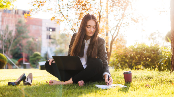 完整的身体尺寸照片年轻女子使用笔记本电脑坐在绿色草地在公园。在线交流。商务人士的生活方式。业务技术。暑假。图片下载
