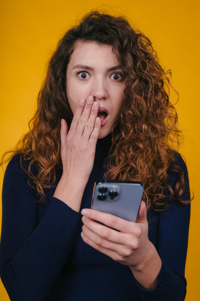 一个美丽的震惊的年轻卷发妇女的形象孤立在黄色的墙壁背景使用手机。图片下载