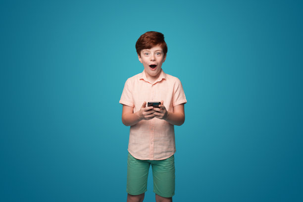兴奋惊讶的红头发男孩拿着手机接收信息与推广孤立在蓝色背景。在线交流。手机通讯。数字技术。图片下载